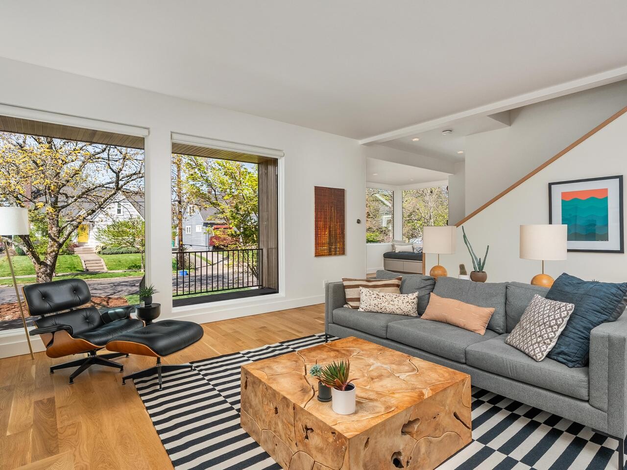 A modern living room with a gray sofa, wooden coffee table, black lounge chair, striped rug, and large windows showing a green outdoor scene. Decorative pillows, lamps, and framed art complete the cozy, bright space.