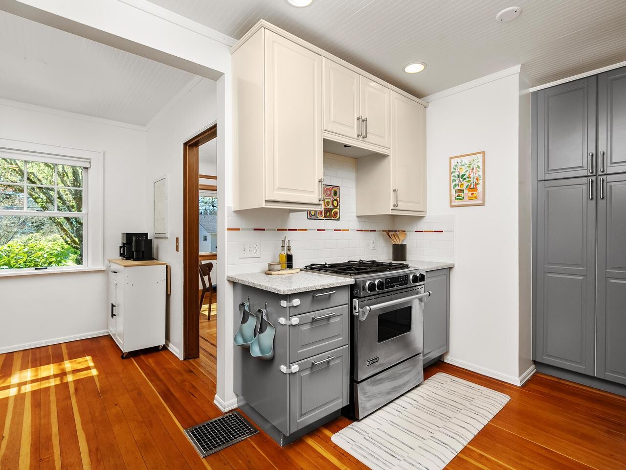 Bright, modern kitchen with white upper cabinets, gray lower cabinets, a gas stove, oven, and hood. There’s a kitchen mat, wooden floor, pantry, small counter with a coffee maker, and a window letting in natural light.
