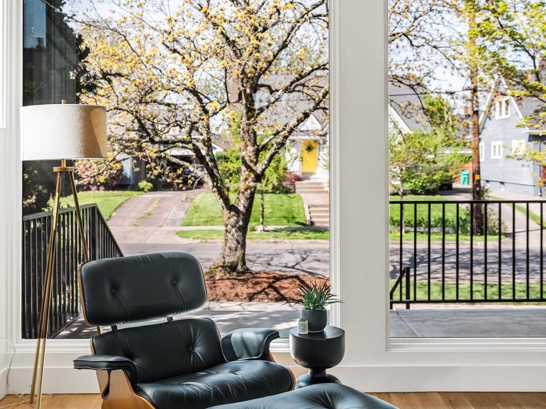 A modern lounge chair with ottoman sits by large windows, offering a view of a tree-lined suburban street. A small side table with a plant and a floor lamp complete the bright, cozy corner on wooden floors.