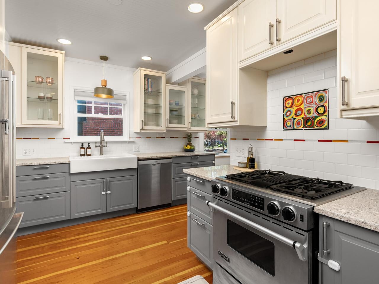 Modern kitchen with white upper cabinets, gray lower cabinets, stainless steel appliances, a farmhouse sink under a window, wood floors, and a colorful abstract tile above the stove.