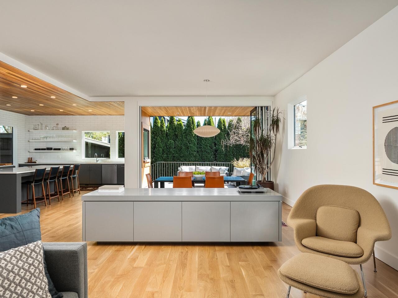 Modern open-concept living space with light wood floors, a cozy chair, gray cabinets, kitchen with barstools, dining area, and glass doors opening to a patio with outdoor seating and greenery.