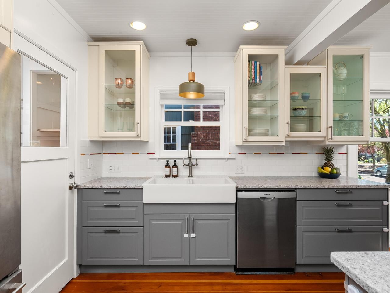 Modern kitchen with gray lower cabinets, white upper cabinets with glass doors, a farmhouse sink, stainless steel dishwasher, wooden floors, and a window above the sink letting in natural light.