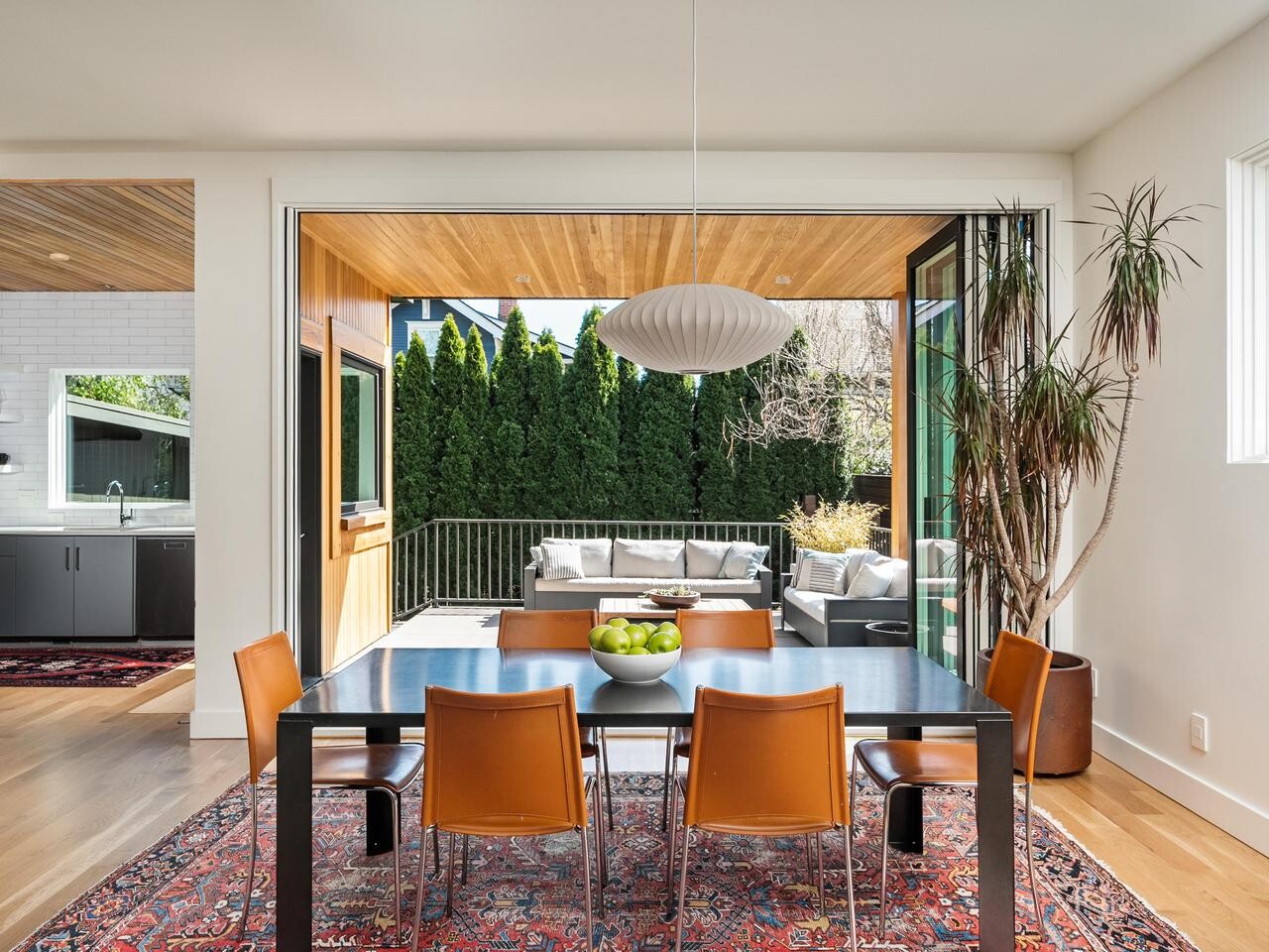 Modern dining area with six orange chairs around a black table on a patterned rug, large window views to a patio with outdoor seating, and adjacent open kitchen with light wood floors and abundant natural light.