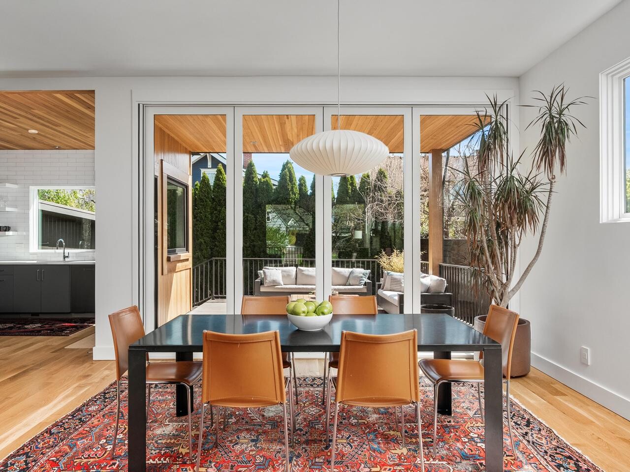 Modern dining room with a black table, six brown chairs, and a red patterned rug. Large windows open to a patio with outdoor seating. A white pendant light hangs above the table, and a tall plant sits by the window.