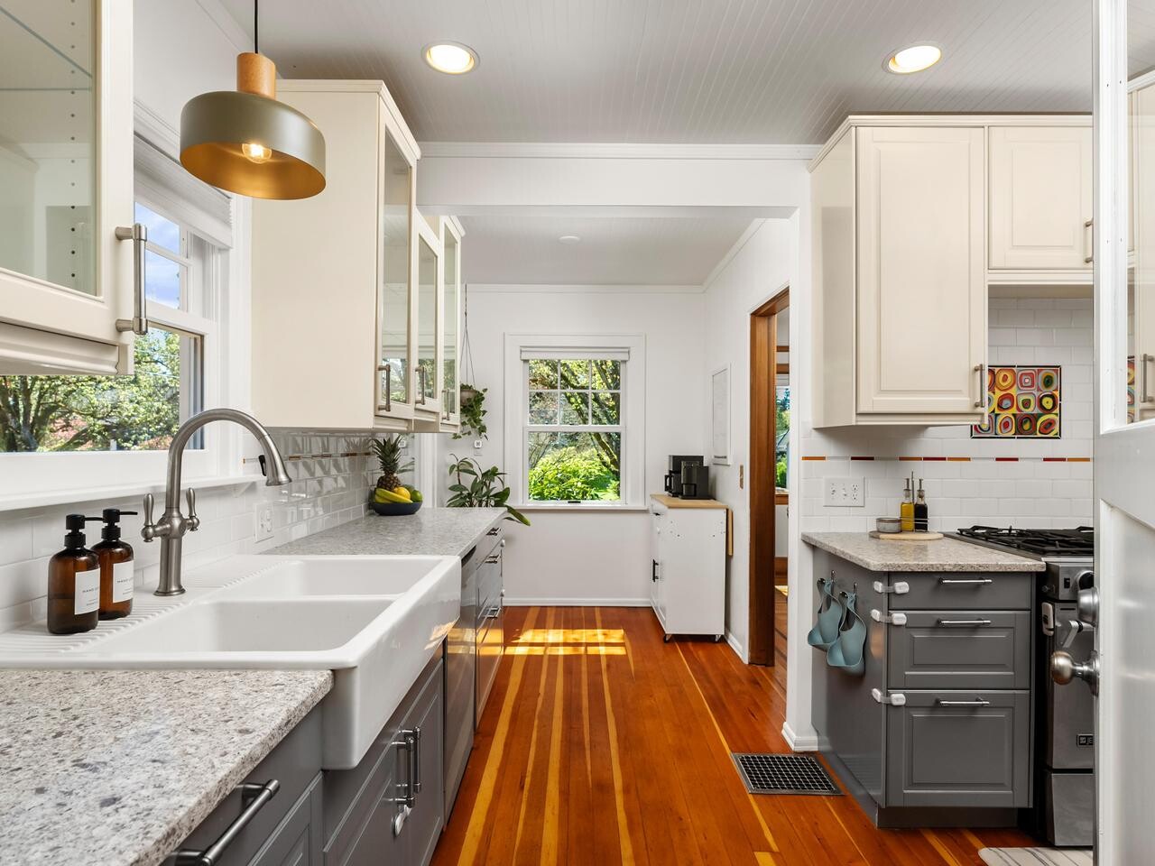Modern kitchen with white and gray cabinets, marble countertops, a farmhouse sink, stainless steel appliances, wooden floors, and large windows letting in natural light. Some plants and soap bottles sit by the window.