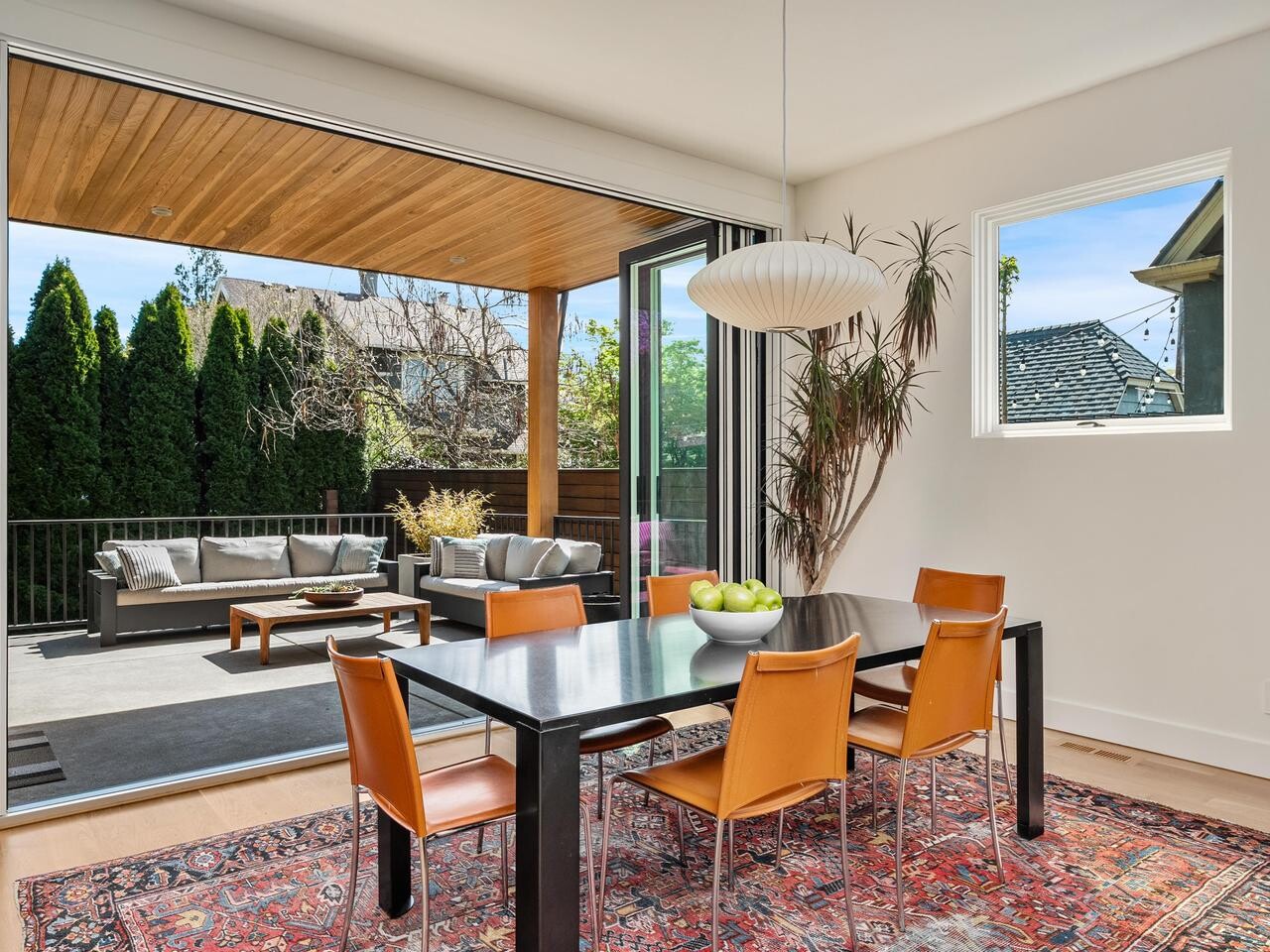 Modern dining room with a black table, orange chairs, and a bowl of apples on a patterned rug, opening to a patio with outdoor seating and greenery. Large windows let in natural light.