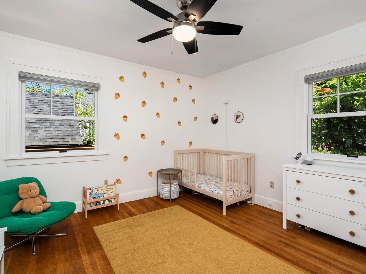 Bright nursery with a beige crib, white dresser, green chair with a teddy bear, and a yellow rug on a wooden floor. Decorative gold dots on one wall and two large windows with outdoor views. Ceiling fan overhead.