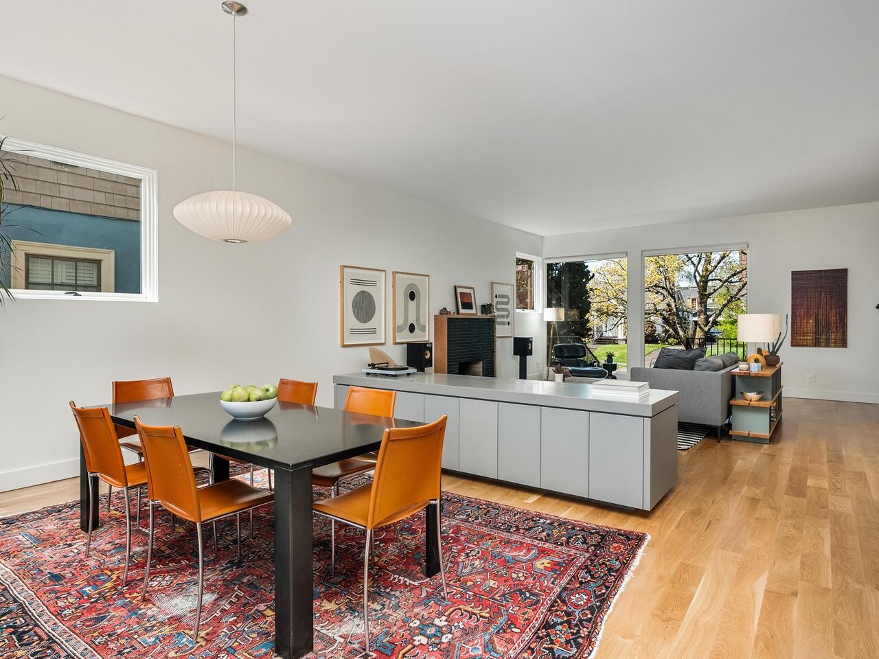 Modern open-concept dining and living room with a black table, six orange chairs on a patterned rug, pendant light, wooden floors, and large windows offering natural light and a view of greenery.