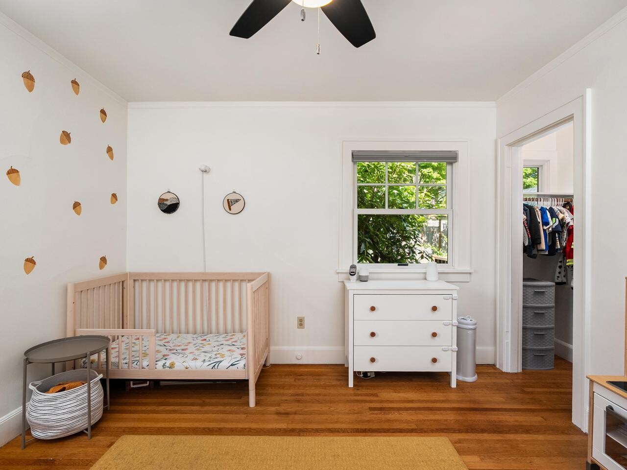 A nursery room with a wooden crib, white dresser with changing pad, acorn decals on the wall, hardwood floor, a window with leafy trees outside, and an open door leading to a closet.