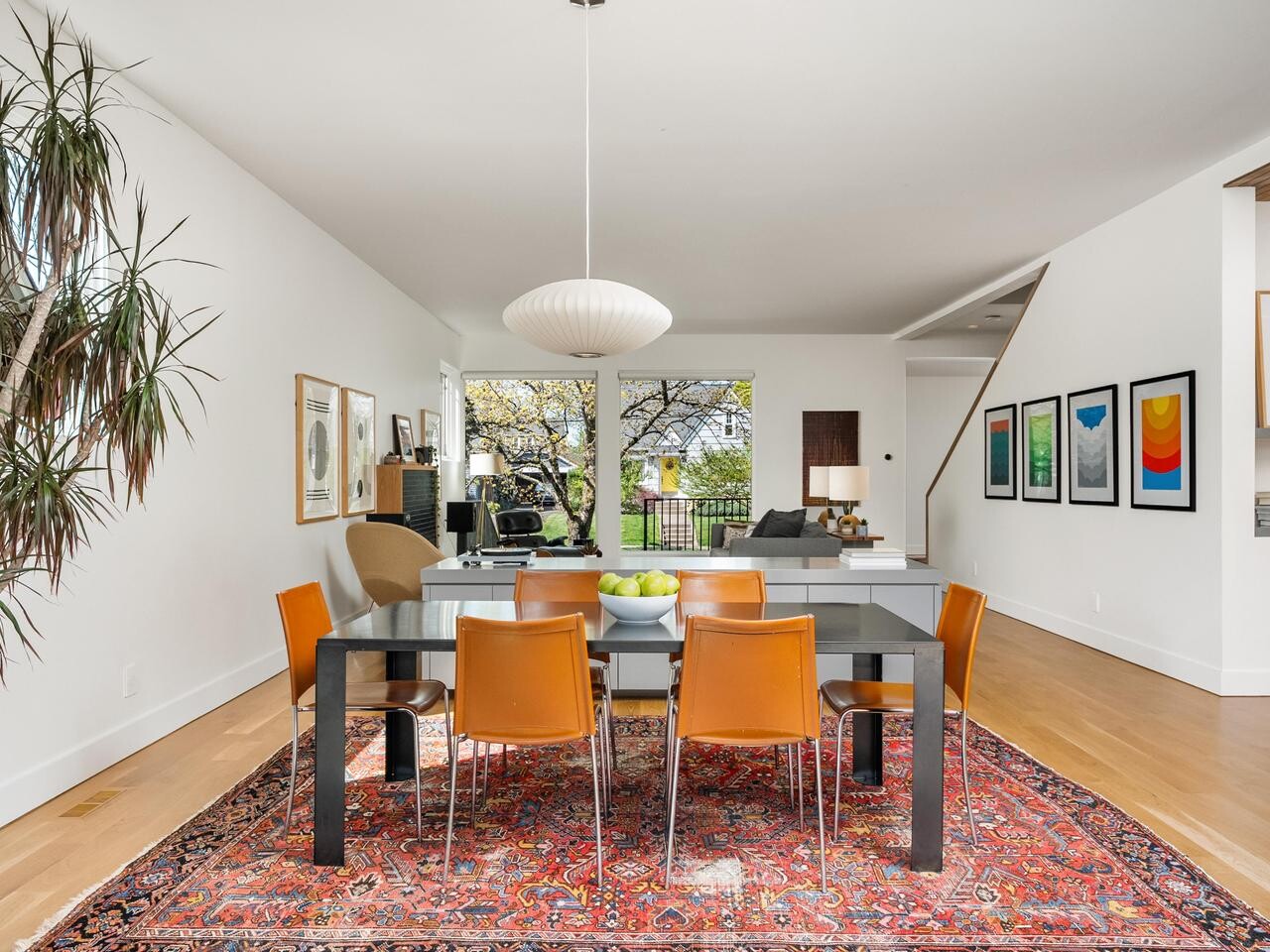 A modern dining room with a black table, six brown chairs, a large red patterned rug, and a round white pendant light. Artworks and plants decorate the space, with living area and large windows in the background.