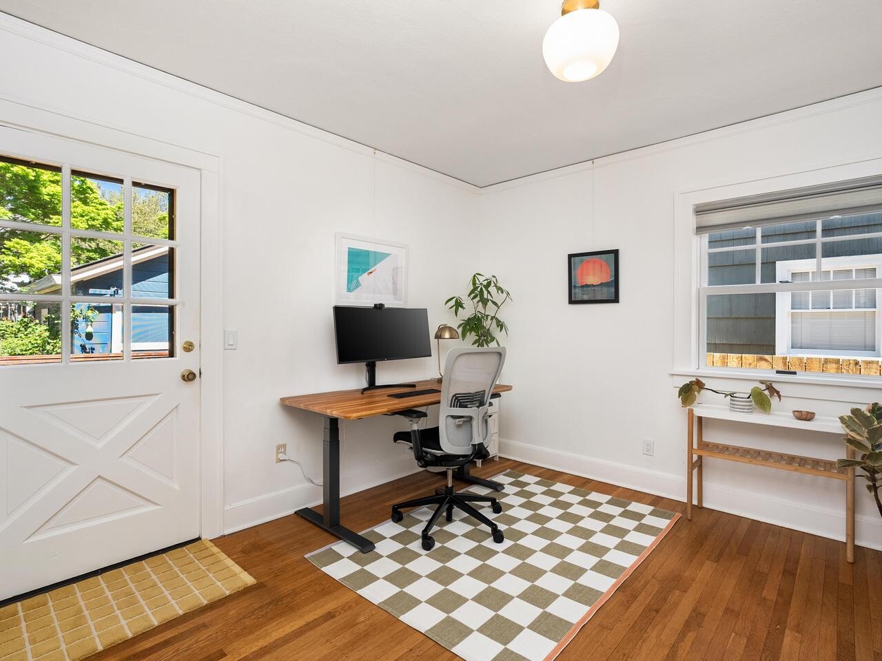 A bright home office with white walls, a wooden floor, a desk with a computer and ergonomic chair, a checkered rug, a plant, a bookshelf, and windows letting in natural light.