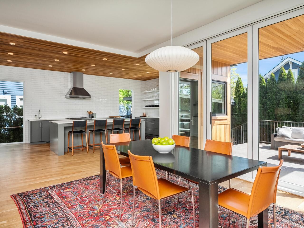Modern dining room with orange chairs around a black table on a red patterned rug, adjacent to an open kitchen with gray cabinets and a breakfast bar. Large glass doors open to a sunny patio with outdoor seating.