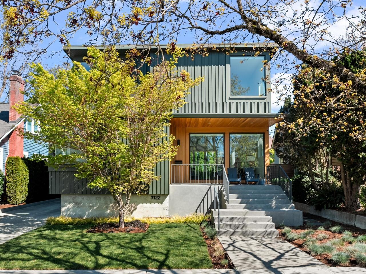 A modern two-story house with gray vertical siding, large windows, and a covered porch. A tree with green leaves stands in front, and the driveway and manicured lawn are visible.