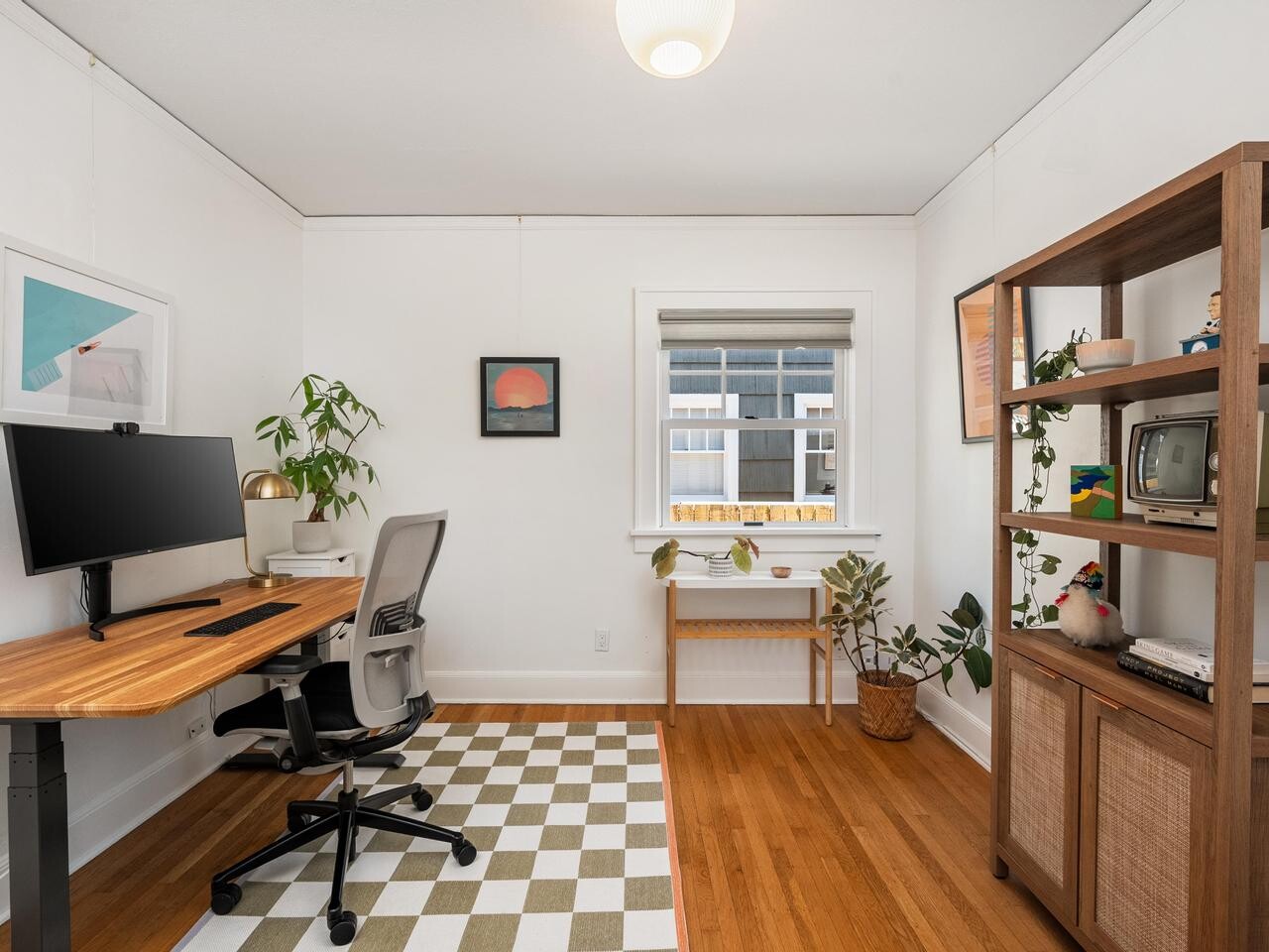A modern home office with a wooden desk, monitor, office chair, potted plants, artwork on white walls, a checkered rug, wooden floor, and a window letting in natural light. A vintage TV and books are on a wooden shelf.
