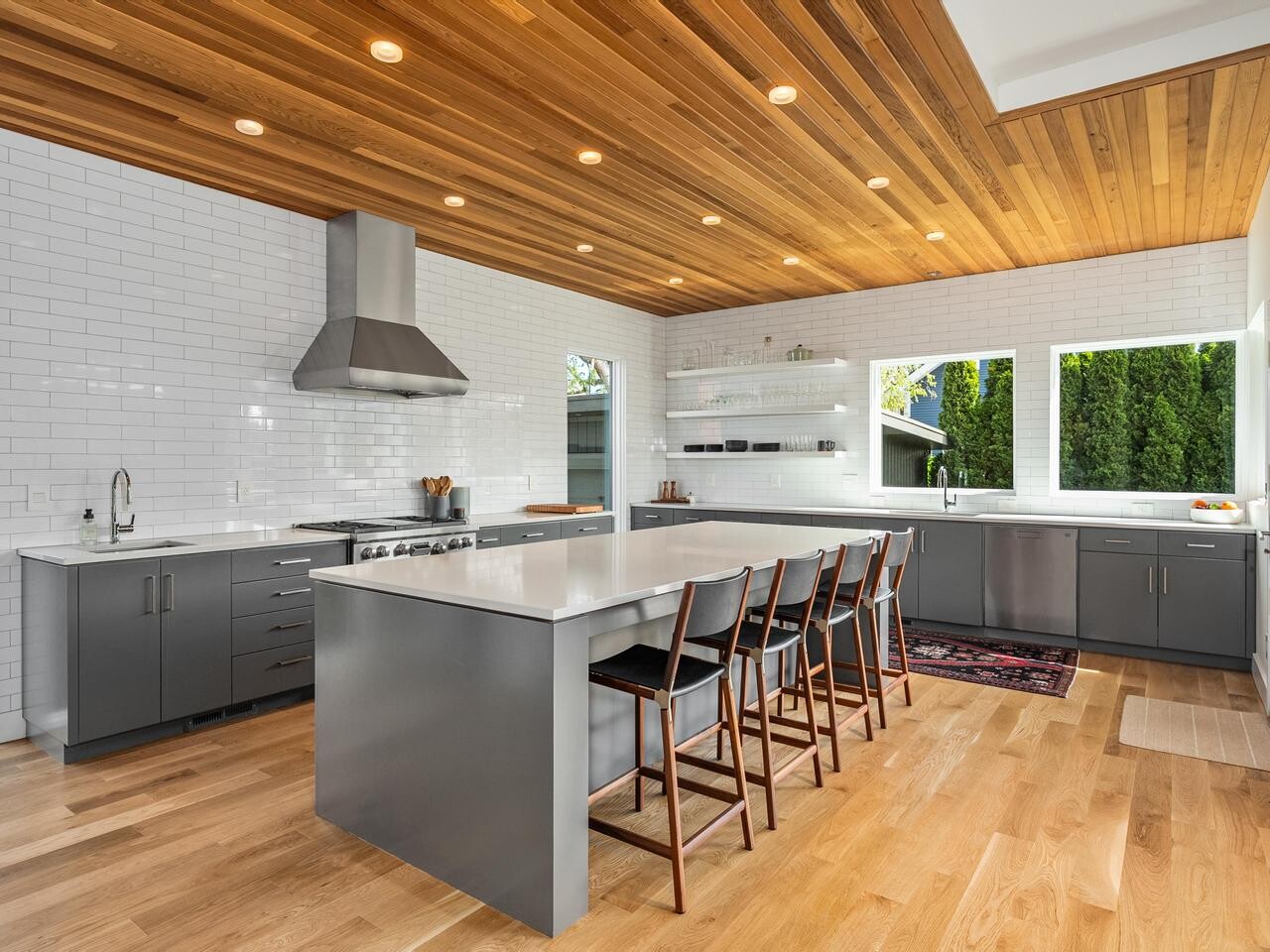 Modern kitchen with sleek gray cabinets, white countertops, and a large island with four wooden barstools. The ceiling is wood-paneled, and the space is well-lit with recessed lights and large windows.