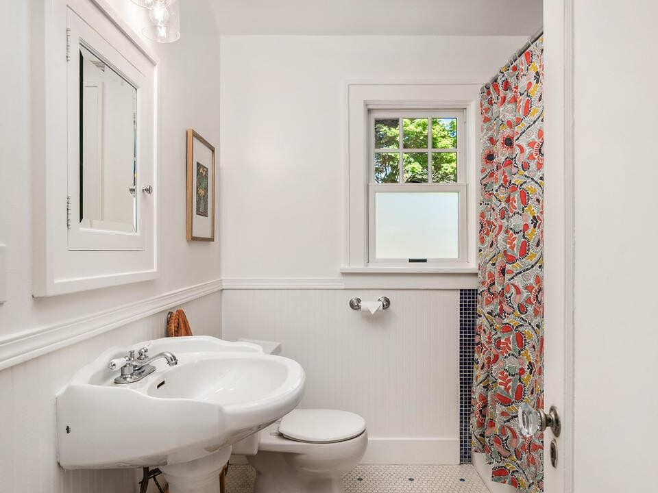 A small, bright bathroom with white walls, pedestal sink, toilet, a window, and a colorful shower curtain. The floor has white hexagonal tiles with blue accents, and there is a three-light fixture above the mirror.