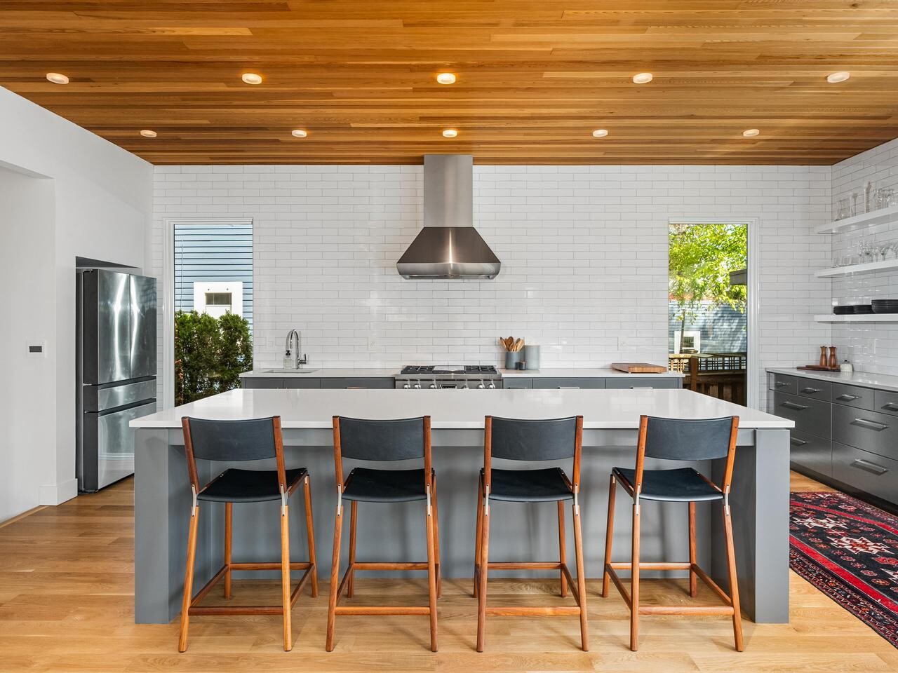 Modern kitchen with a wooden ceiling, white subway tile walls, a large gray island with four black chairs, stainless steel appliances, open shelving, and a colorful rug on hardwood floors. Natural light comes through two windows.