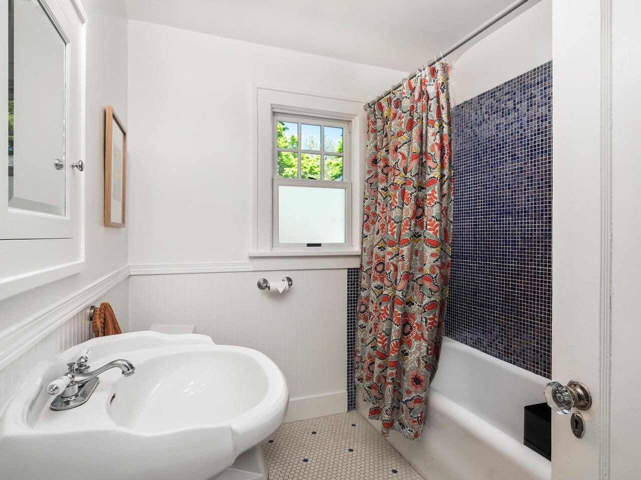 A small, bright bathroom with a white sink, toilet, and tub. The shower has a colorful patterned curtain and dark blue tile walls. A window lets in natural light above the toilet.