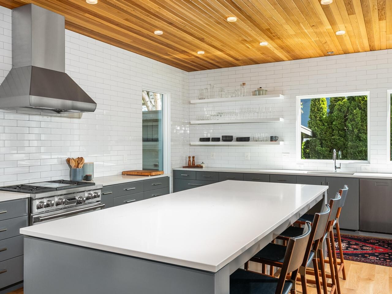 Modern kitchen with gray cabinets, a large white island, stainless steel range hood, open shelves with glassware, white subway tile backsplash, wood-paneled ceiling, and large windows overlooking greenery.