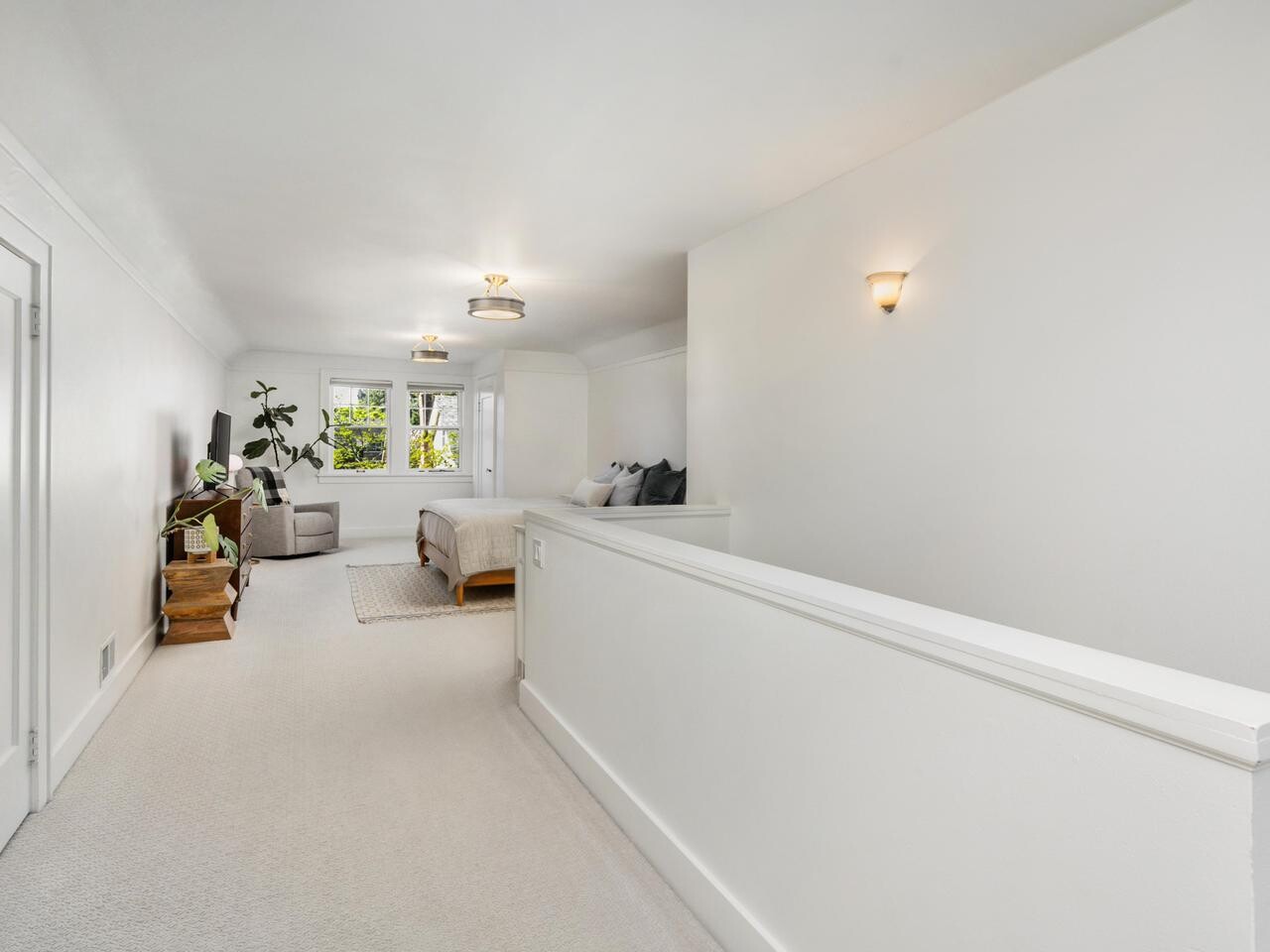 A bright, white hallway leads to a spacious bedroom with a large bed, a sofa, plants, wooden furniture, and windows letting in natural light. The space is modern and minimalistic.