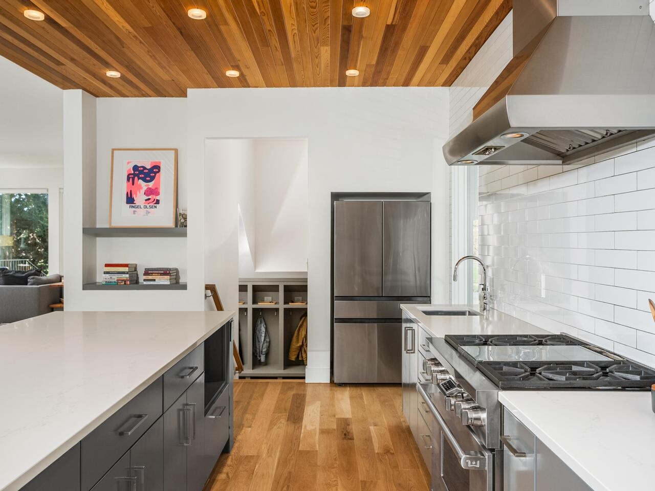 Modern kitchen with stainless steel appliances, white countertops, dark lower cabinets, a wood ceiling, and white subway tile backsplash. An open area with a small coat nook and a framed picture is visible in the background.