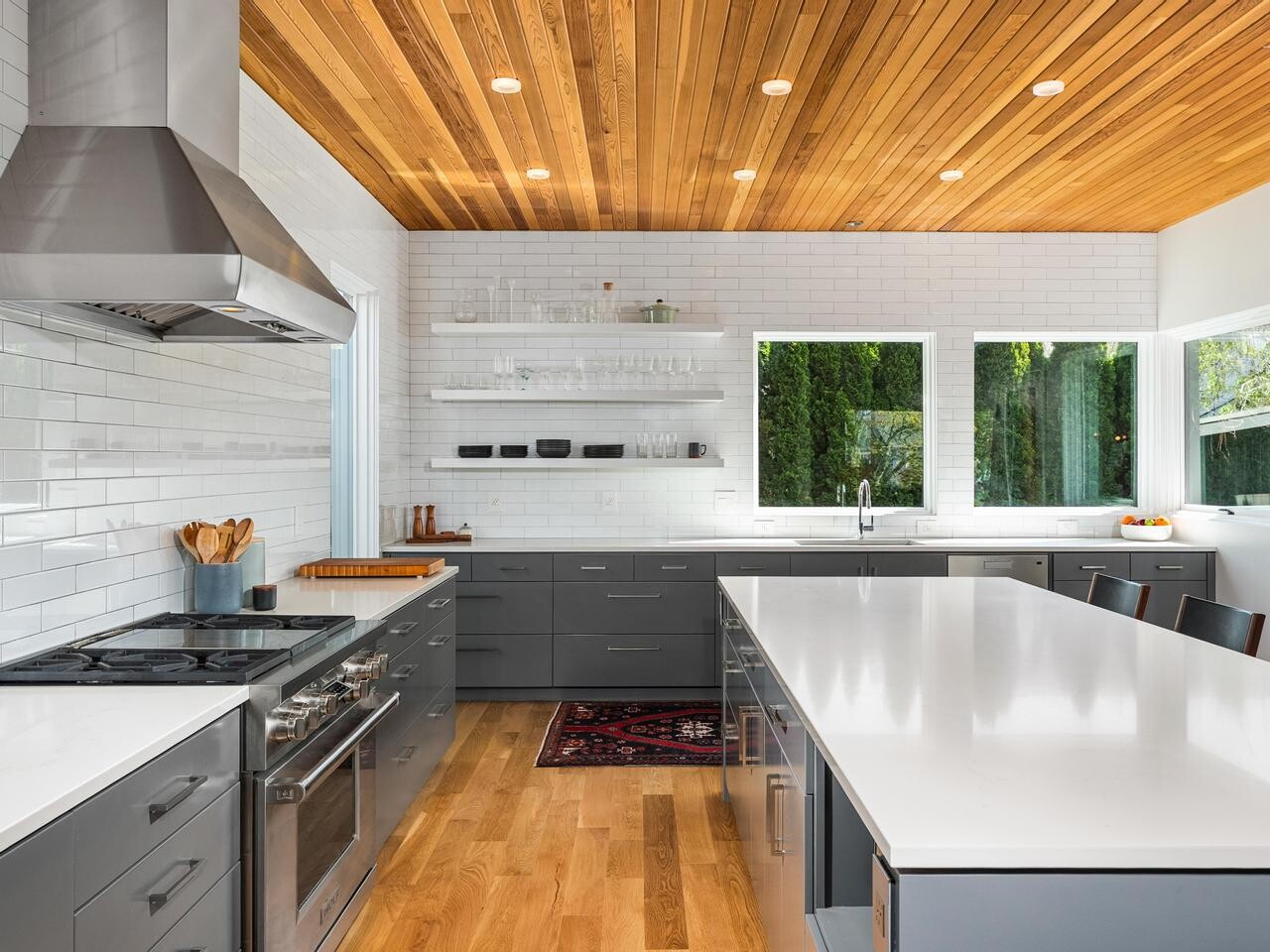 Modern kitchen with gray cabinets, stainless steel appliances, white countertops, a long island with barstools, wooden ceiling, white subway tile backsplash, open shelves, and large windows providing natural light.