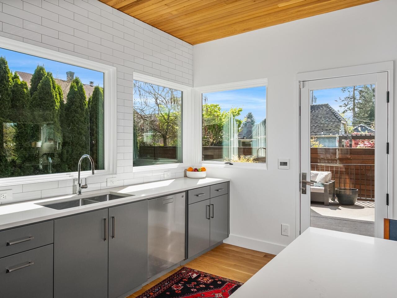 Modern kitchen with gray cabinets, white countertops, subway tile backsplash, and large windows letting in natural light. A bowl of fruit sits on the counter, and a glass door leads to an outdoor patio.