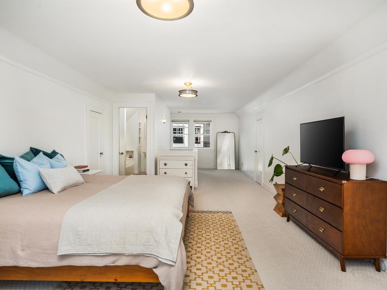 Bright, spacious bedroom with a large bed, blue and beige pillows, a patterned rug, dresser with TV, potted plant, floor mirror, and windows letting in natural light. Walls and carpet are light-colored.