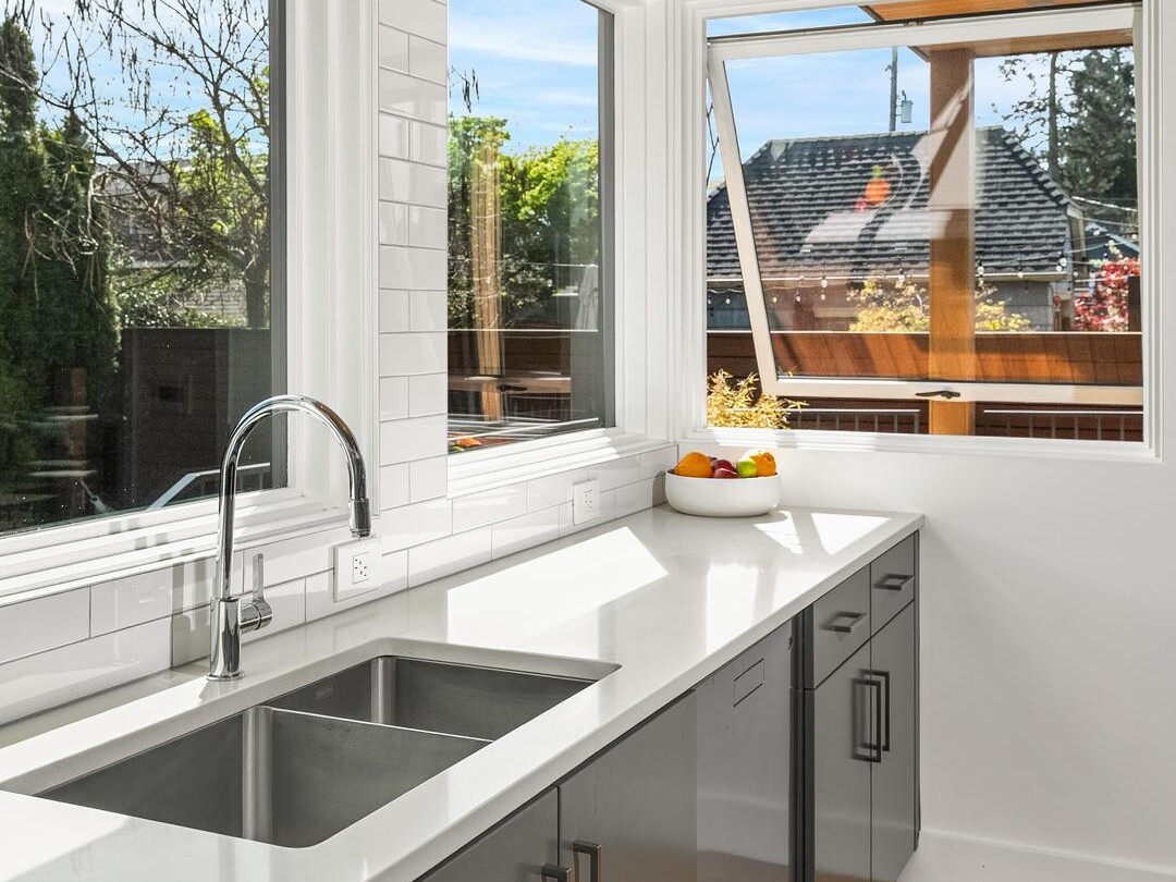 Modern kitchen with gray cabinets, white countertops, double sink, and large windows letting in natural light. A bowl of fruit sits on the counter near an open window with views of greenery and neighboring houses outside.