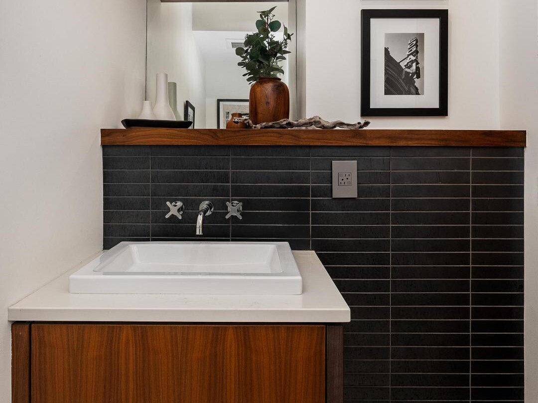 Modern bathroom with a wood vanity, white sink, black tile backsplash, open wood shelves with decor, and two black-and-white framed photos on a white wall. Geometric black and white floor tiles are visible.