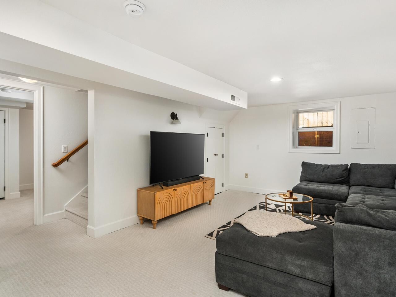 A bright, carpeted living room with white walls, a large gray sectional sofa, a wooden TV stand with a flat-screen TV, and a small round table. A window lets in natural light, and stairs lead to another area.
