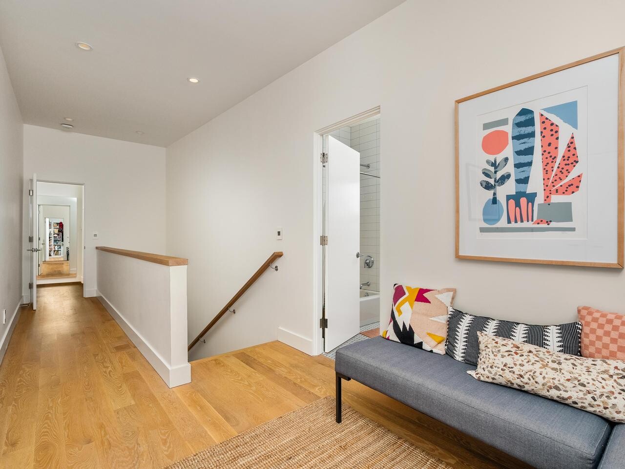A modern hallway with light wood flooring, white walls, a bench with colorful cushions, a woven rug, and abstract artwork above the bench. Open doors lead to other rooms and a staircase descends to the left.