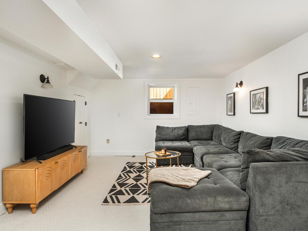 A cozy living room with a large gray sectional sofa, a black-and-white patterned rug, a light wood TV stand with a flat-screen TV, wall art, and a small round coffee table under a window.