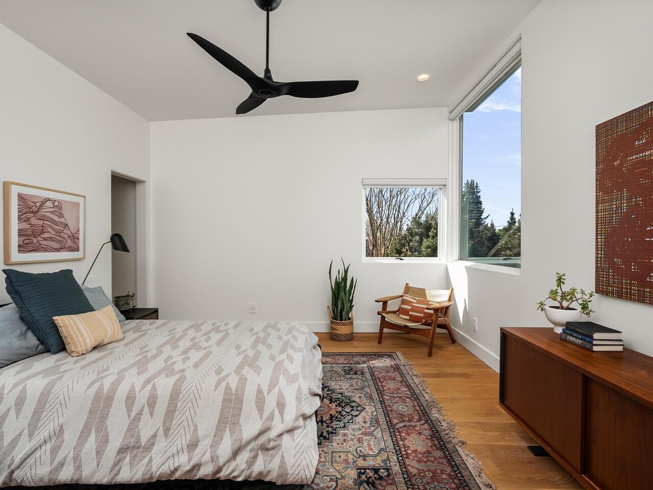 A modern bedroom with a patterned bed, pillows, a colorful rug, wooden furniture, a potted plant, artwork on the walls, a ceiling fan, and large windows letting in natural light.