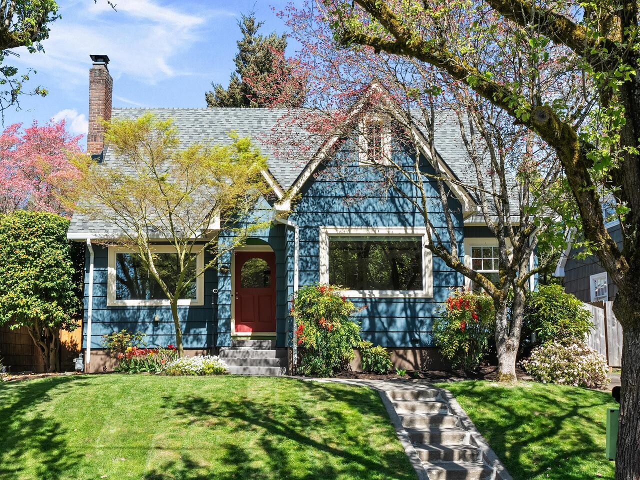 A charming blue house with a red front door, large windows, and a gabled roof. The front yard features green grass, blossoming trees, and shrubs, with a stone walkway leading to the entrance.