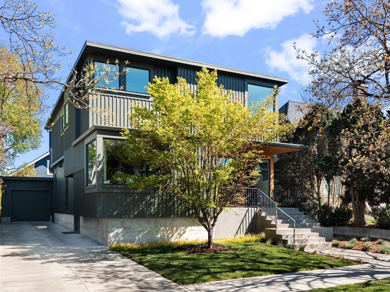 Modern two-story house with dark green vertical siding, large windows, and a front porch. A tree with green leaves stands in the yard, and a concrete driveway and steps lead to the entrance. Blue sky and scattered clouds above.