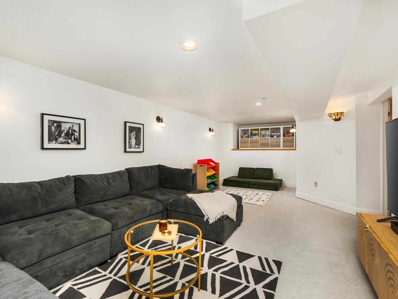 A cozy basement room with a large dark sectional sofa, round glass coffee table, geometric rug, wall art, children’s play area with toys, and a TV on a wooden stand against white walls with recessed lighting.