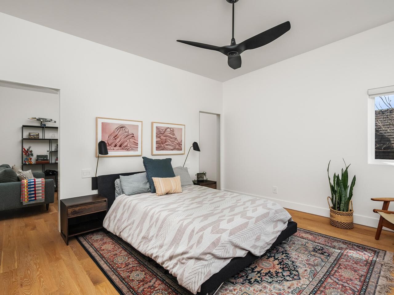 A modern bedroom with a patterned area rug, bed with geometric bedding, two framed artworks above the headboard, and wall-mounted lamps. A potted snake plant and a wooden chair sit near a window. Adjoining room visible.