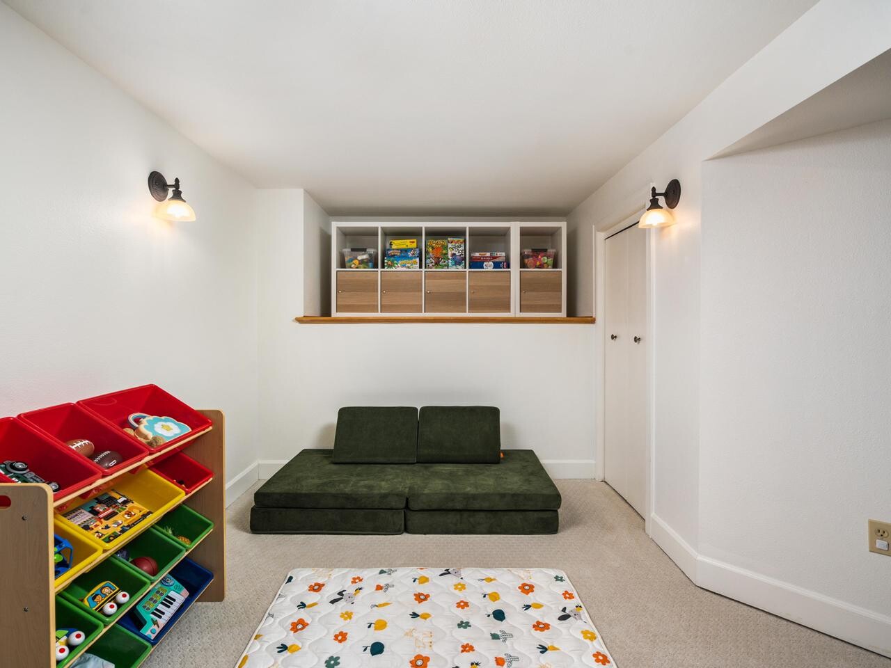 A bright playroom with a green couch, a colorful toy organizer, white walls, built-in shelves holding board games, and a soft playmat with animal patterns on the floor.