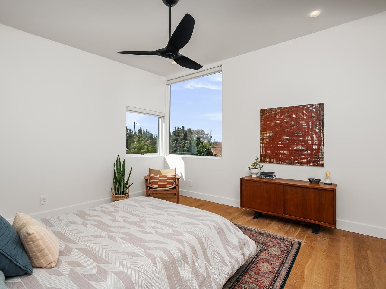 Modern bedroom with white walls, large window, wooden floor, mid-century dresser, abstract red art, potted plant, armchair, ceiling fan, and a bed with geometric-patterned bedding and blue accent pillows.