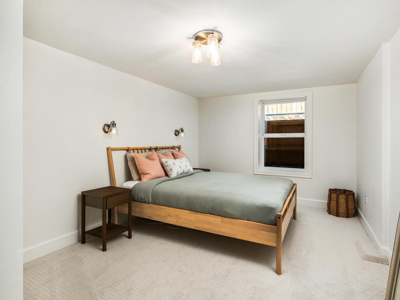 A minimalist bedroom with a wooden bed frame, green bedding, two wall-mounted reading lights, a small bedside table, a wicker basket, a large mirror, and a window letting in natural light.
