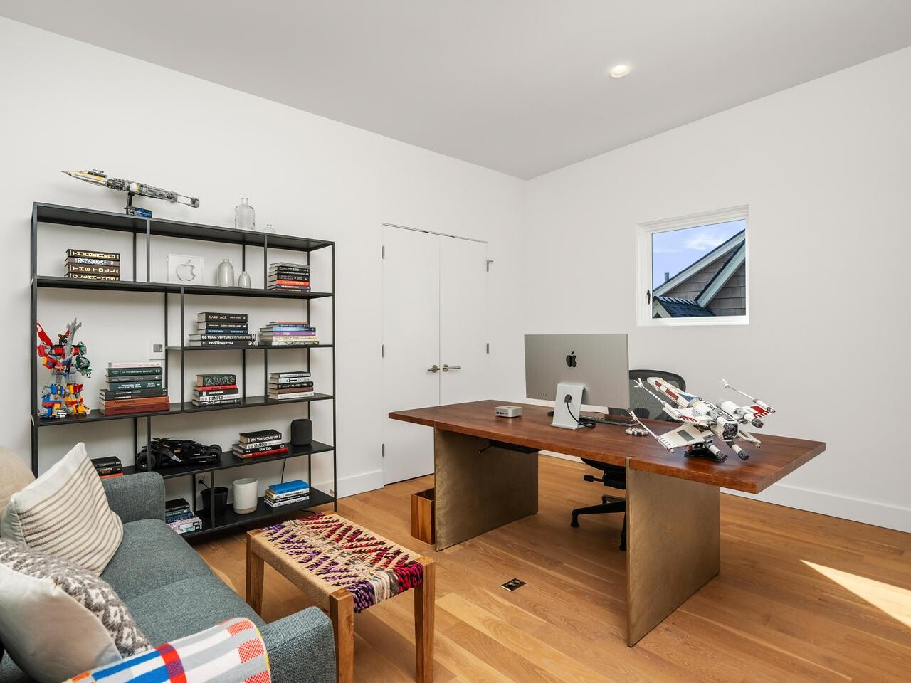 A modern home office with a wooden desk, large monitor, model spaceships, bookshelf filled with books and collectibles, gray sofa with patterned pillow and blanket, and a small window letting in natural light.
