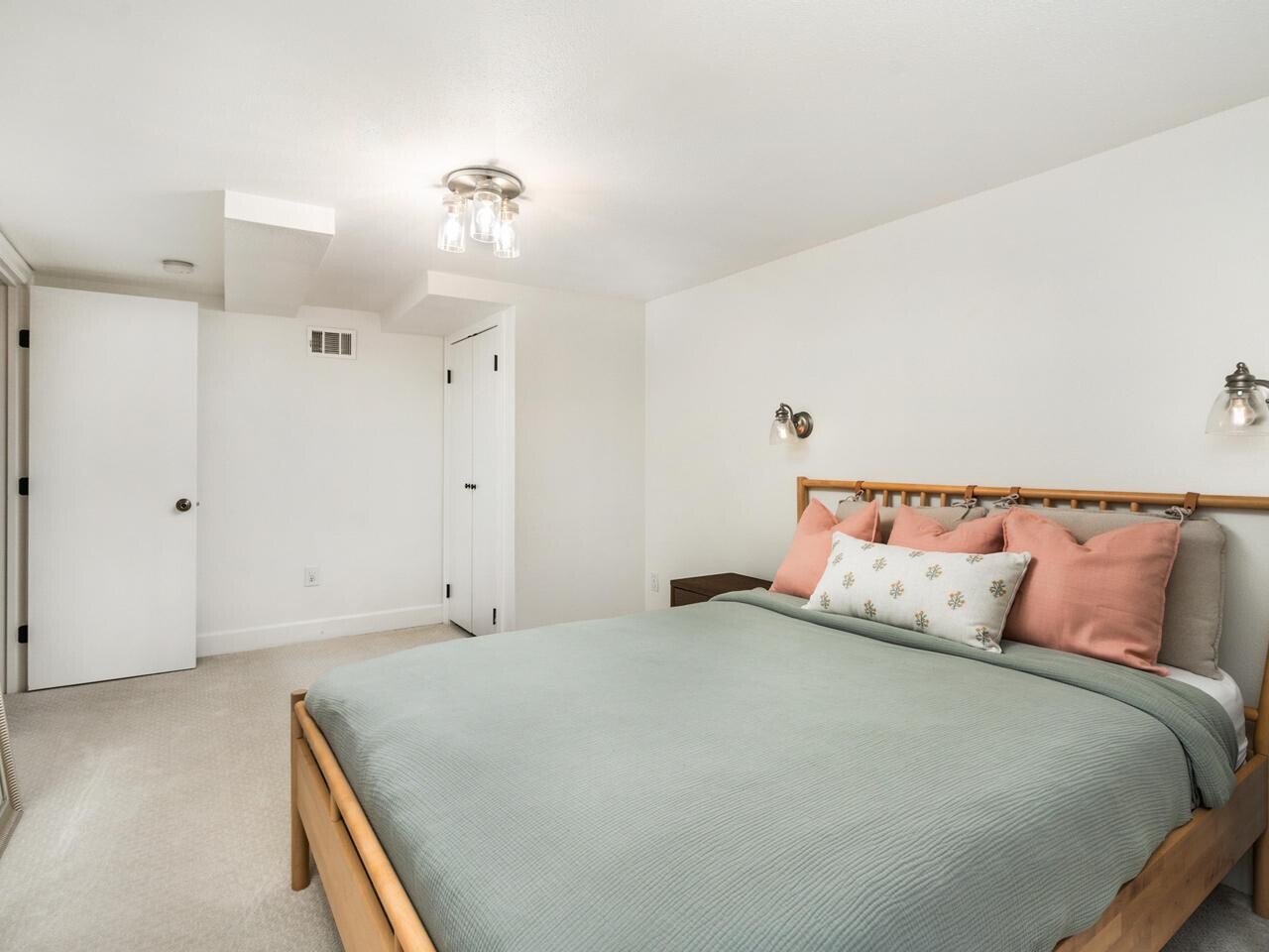 A bright, minimal bedroom with white walls, beige carpet, a wooden bed frame, pastel bedding, wall-mounted lamps, and a large mirror resting against the wall near a closed door.
