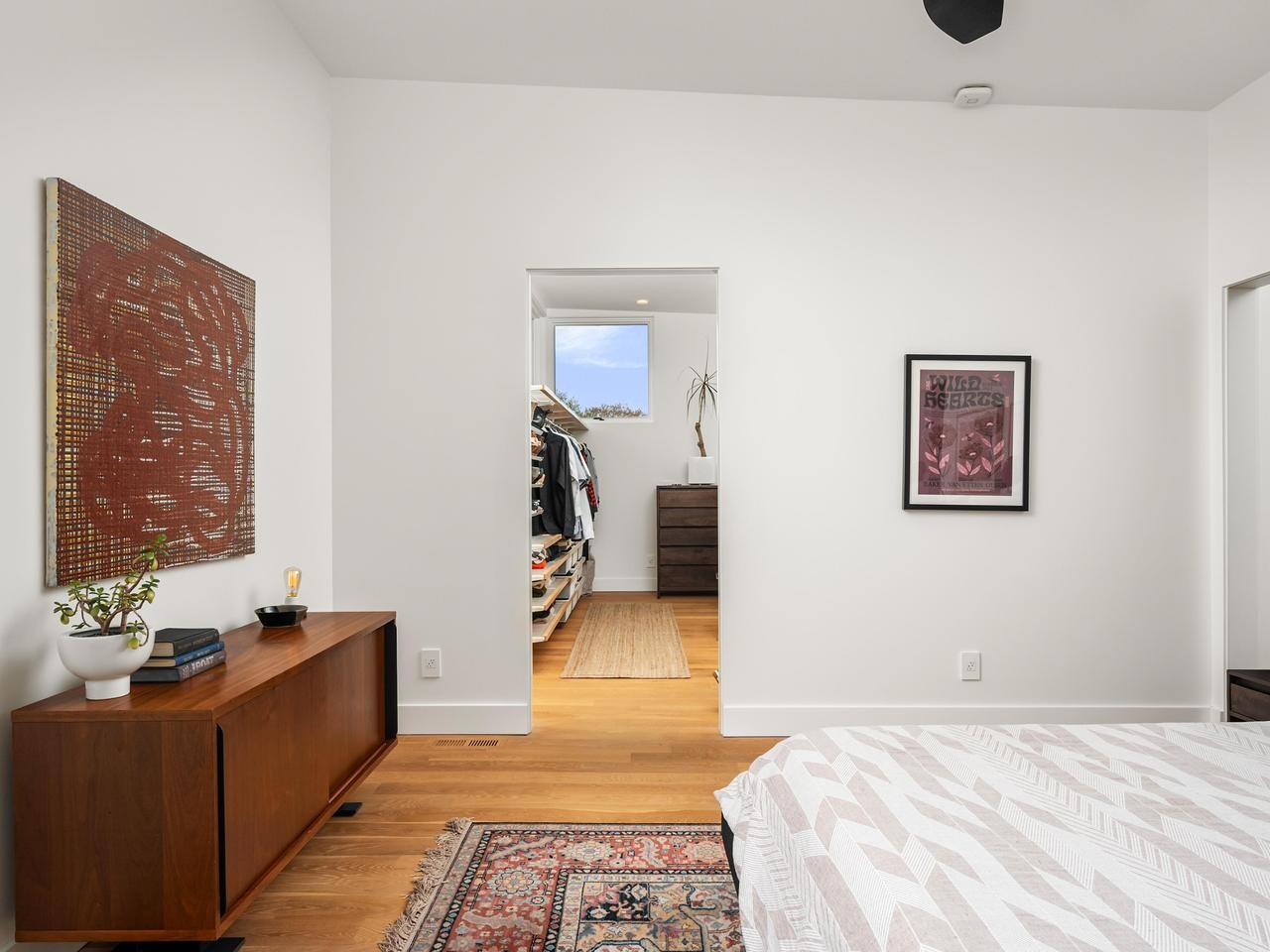 A modern bedroom with white walls, wooden floors, a bed with a patterned blanket, a wooden sideboard with decor, art on the walls, and an open doorway leading to a walk-in closet with clothes and a window.
