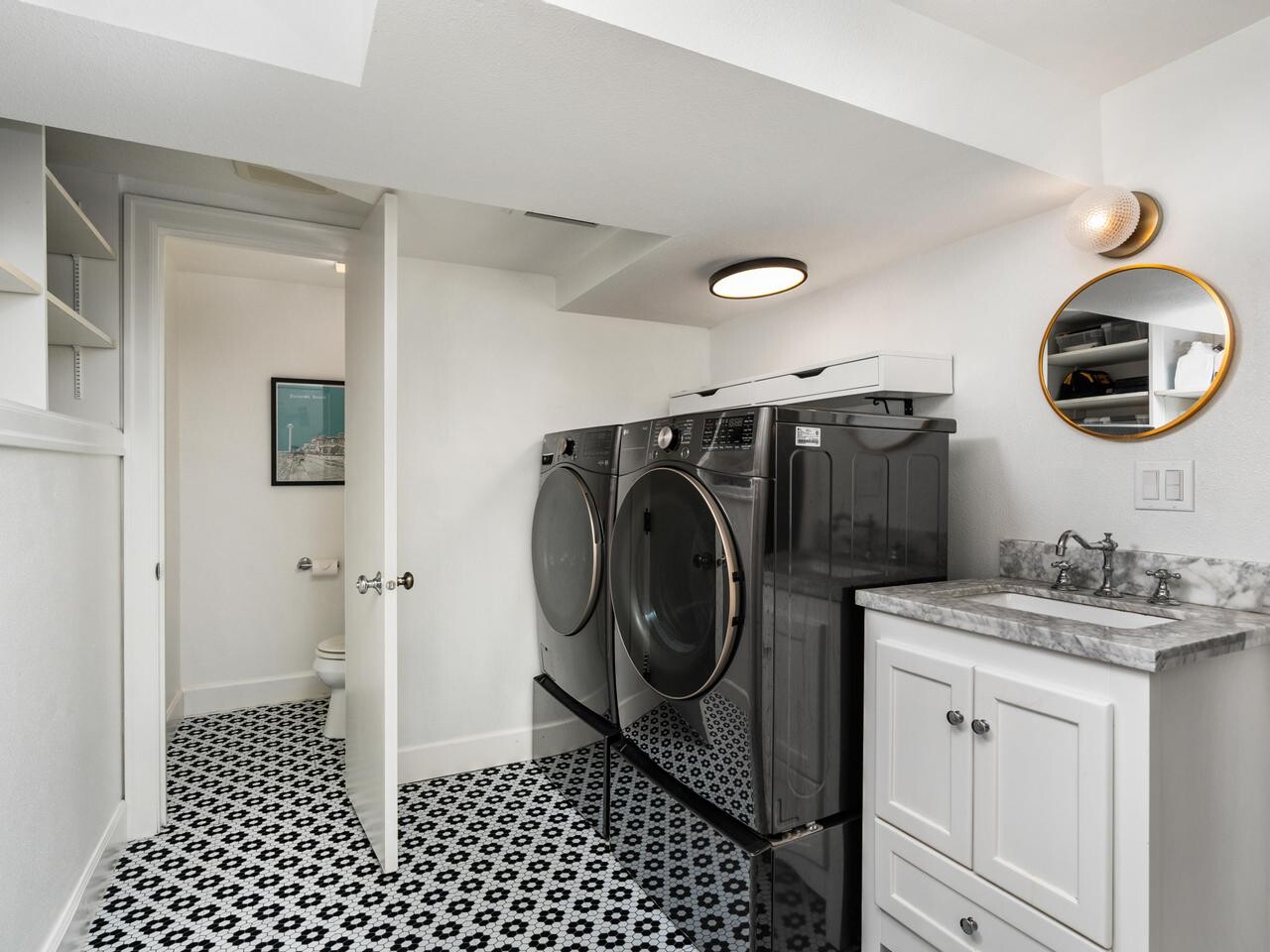 Modern laundry room with black front-load washer and dryer, white cabinets with marble countertop, round mirror, patterned black and white tile floor, and open door leading to a small bathroom.