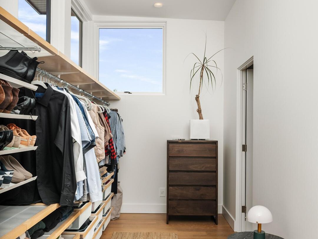 A modern walk-in closet with open shelves and hanging clothes on the left, a woven rug on wooden floors, a small round side table with a lamp, and a dresser with a tall plant near a window at the end.