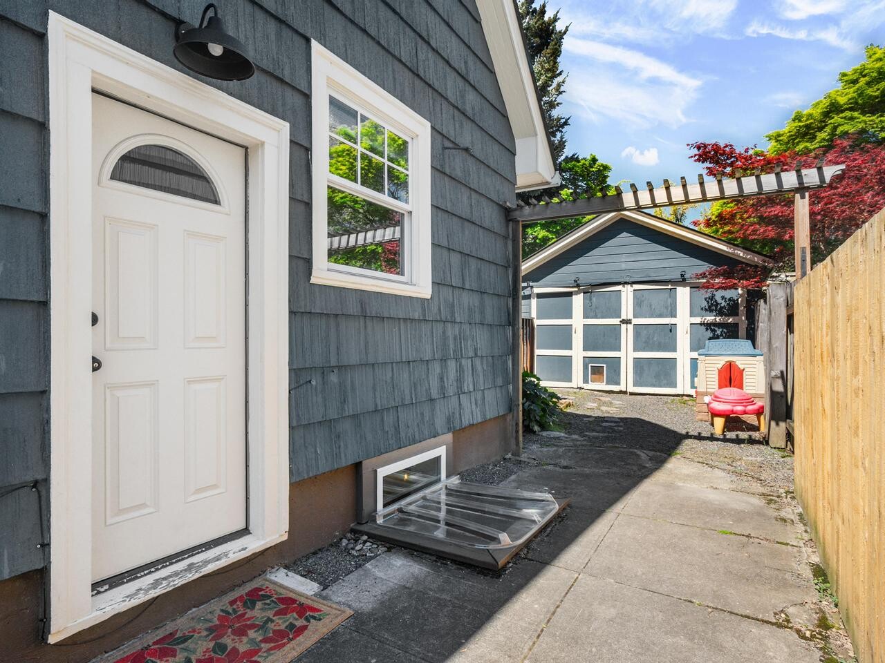 A gray house with a white door and window sits next to a fenced yard. A sidewalk leads to a detached garage. There’s a red chair by a wooden pergola and green trees under a blue sky.