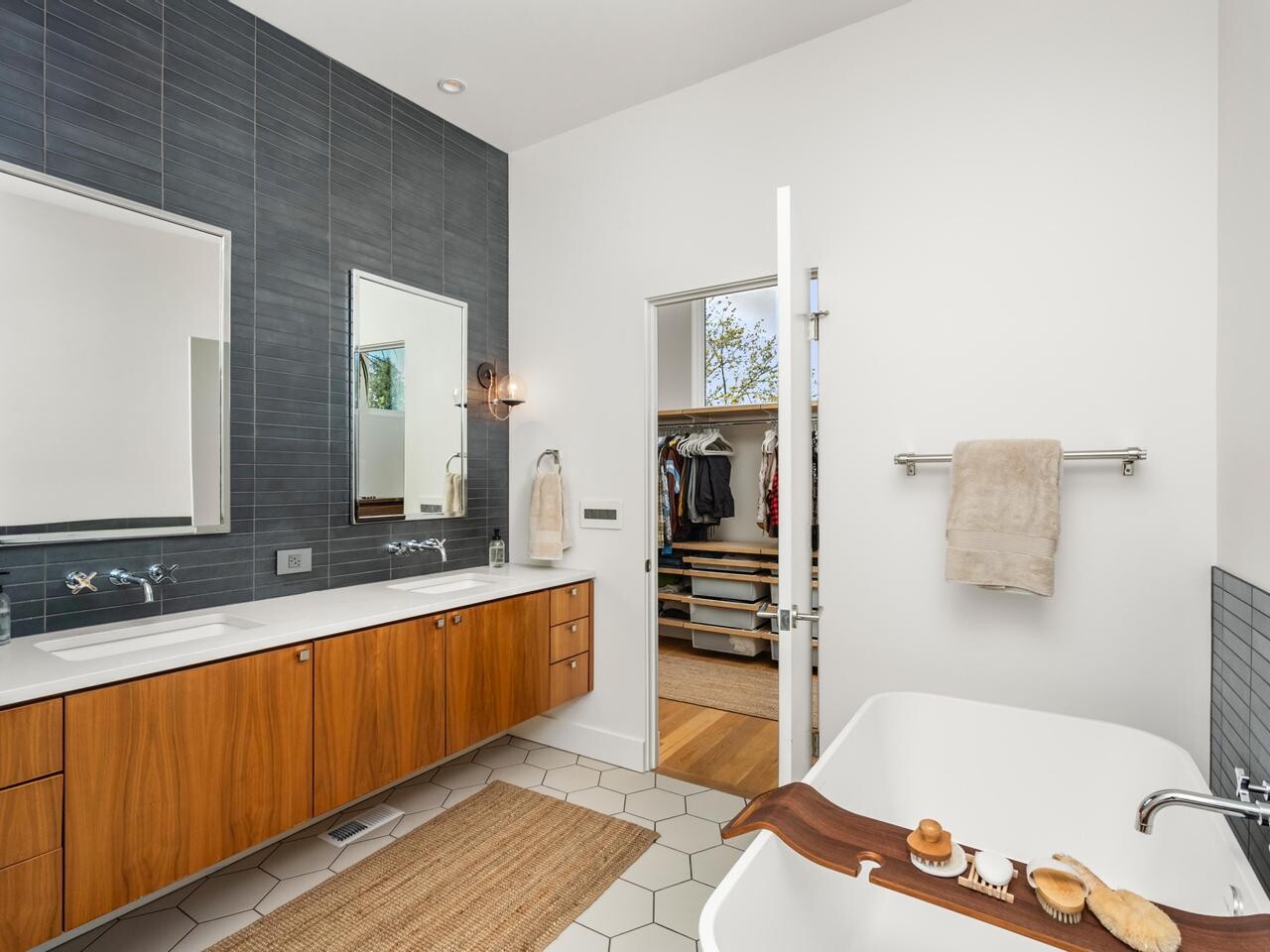 Modern bathroom with a double sink vanity, wood cabinets, two mirrors, a freestanding tub with a wooden tray, hexagonal tile floor, gray tiled wall, and a walk-in closet visible through an open door.