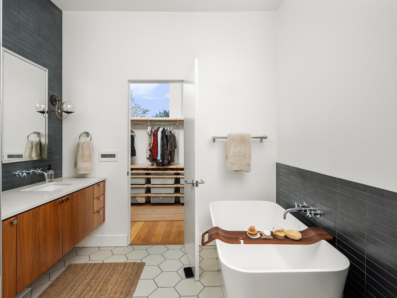 Modern bathroom with white walls, wooden vanity, large mirror, hexagonal tile floor, and a freestanding bathtub. A tray with towels and soap rests on the tub. Door leads to a walk-in closet with shelves and hanging clothes.