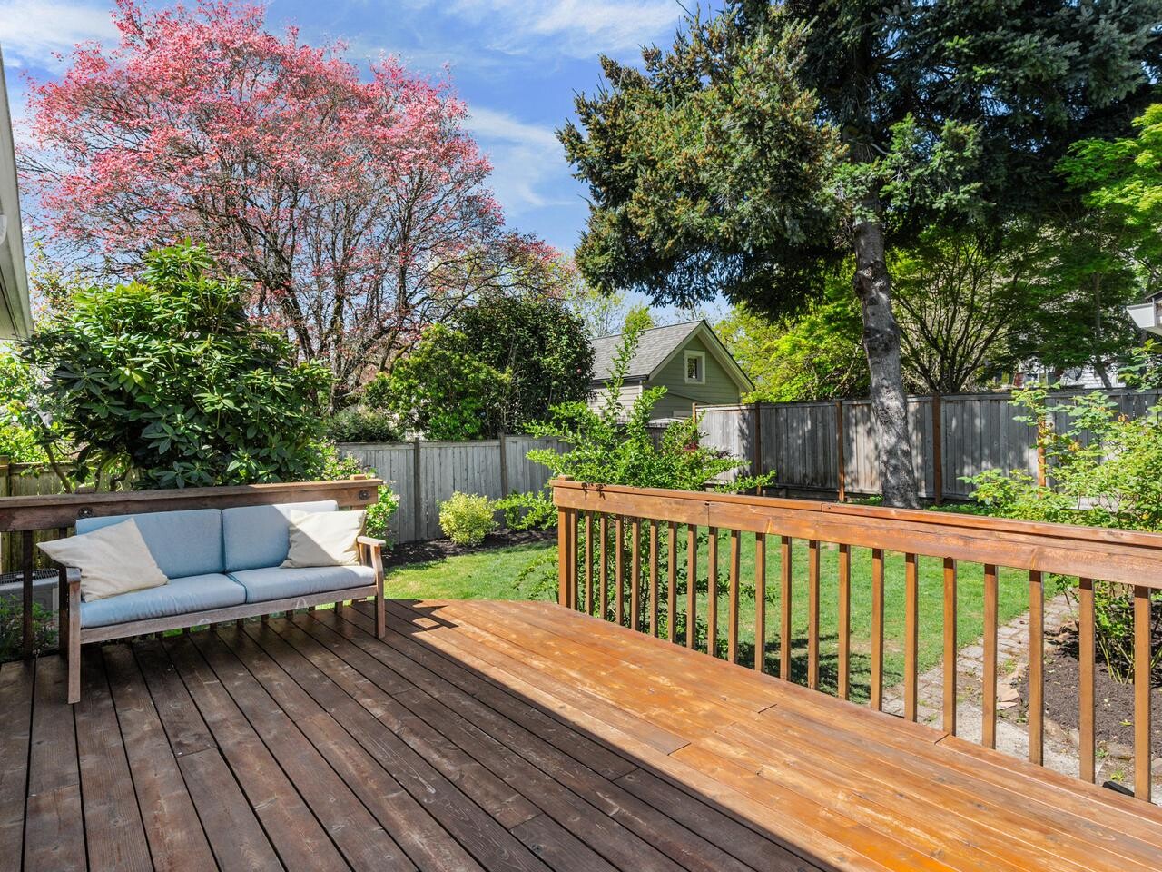 A wooden deck with a cushioned outdoor bench overlooks a fenced backyard with green grass, trees, shrubs, and a house visible in the background under a blue sky with clouds.